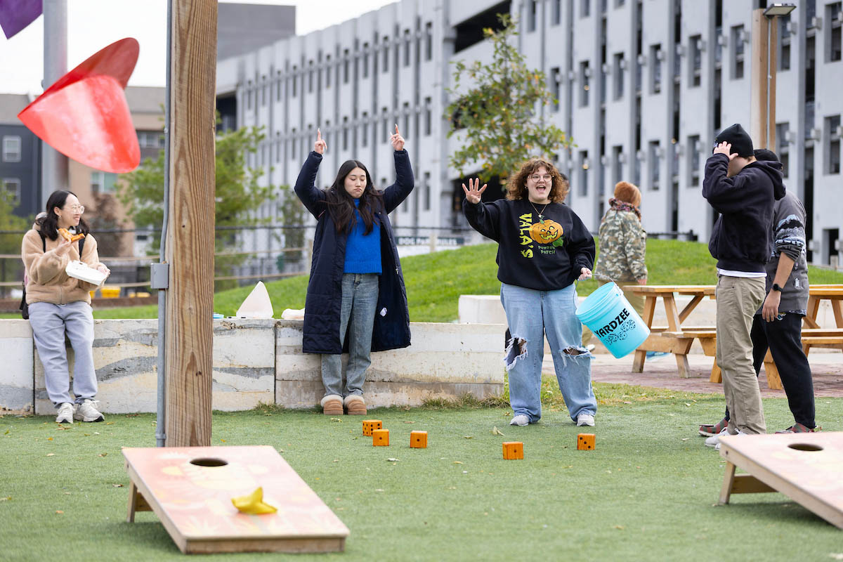 A group of students is gathered outdoors, engaged in games. Two individuals are celebrating with raised hands while others are positioned near game setups. Nearby, a picnic area with wooden tables is visible.