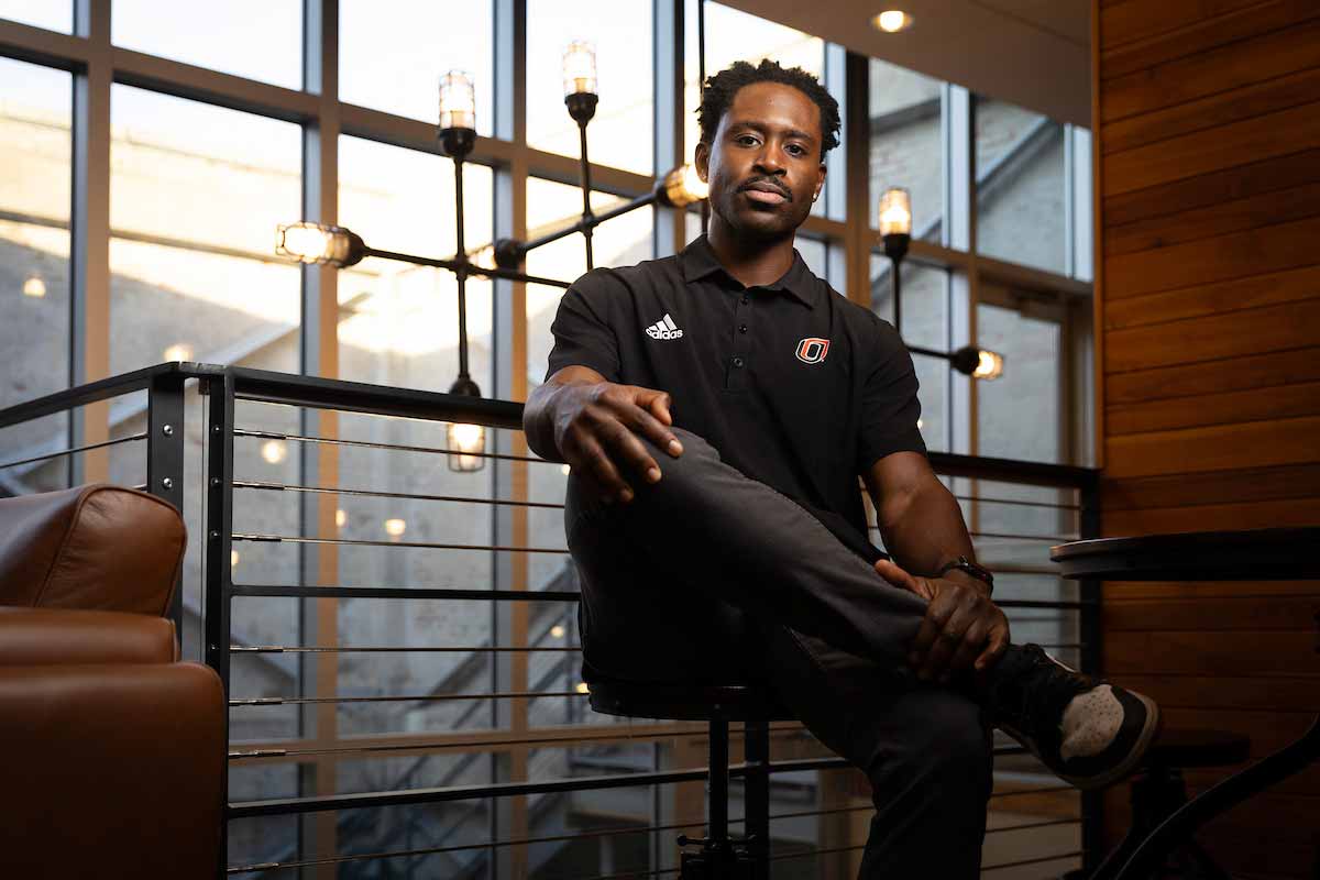 Fabio Gbetanou, a recruiter for UNO, sits confidently in a modern, well-lit indoor space with large windows and industrial-style lighting, wearing a black UNO polo shirt and looking toward the camera. 