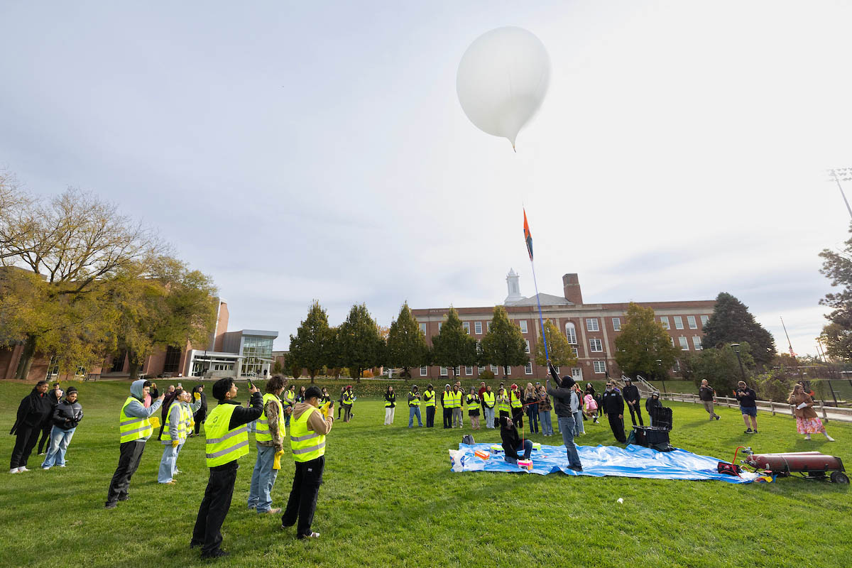 A group of people in bright vests are gathered outdoors on a grassy area, releasing a large white balloon into the sky. Some individuals are holding the balloon while others observe from a distance.