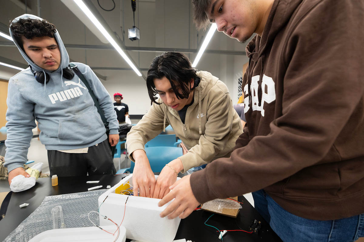 Three students are gathered around a table in a classroom. One student is focused on adjusting components inside a white device, while the others observe closely. The environment features modern furnishings and bright lighting, indicating an educational setting.