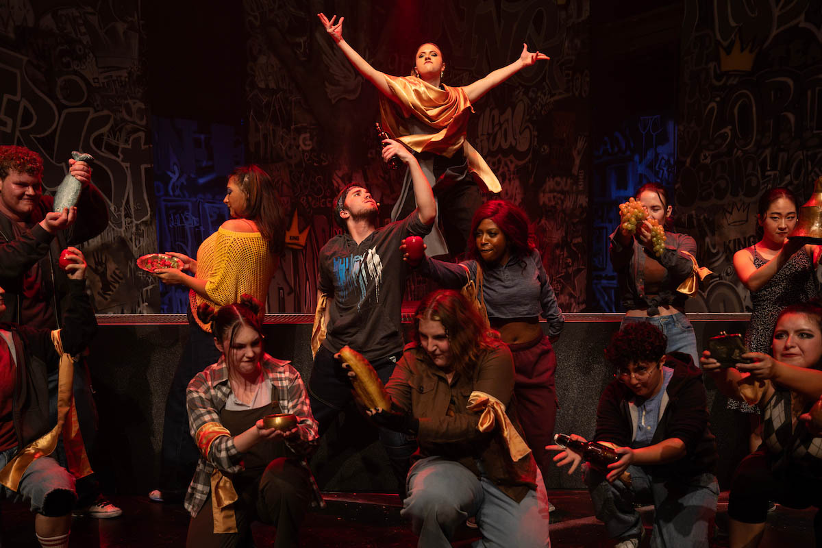 A dynamic theater production scene featuring a diverse group of actors. One performer in a gold costume is elevated above the others, who are arranged around her in various poses. The backdrop includes graffiti-like designs, contributing to the energetic atmosphere of the performance.