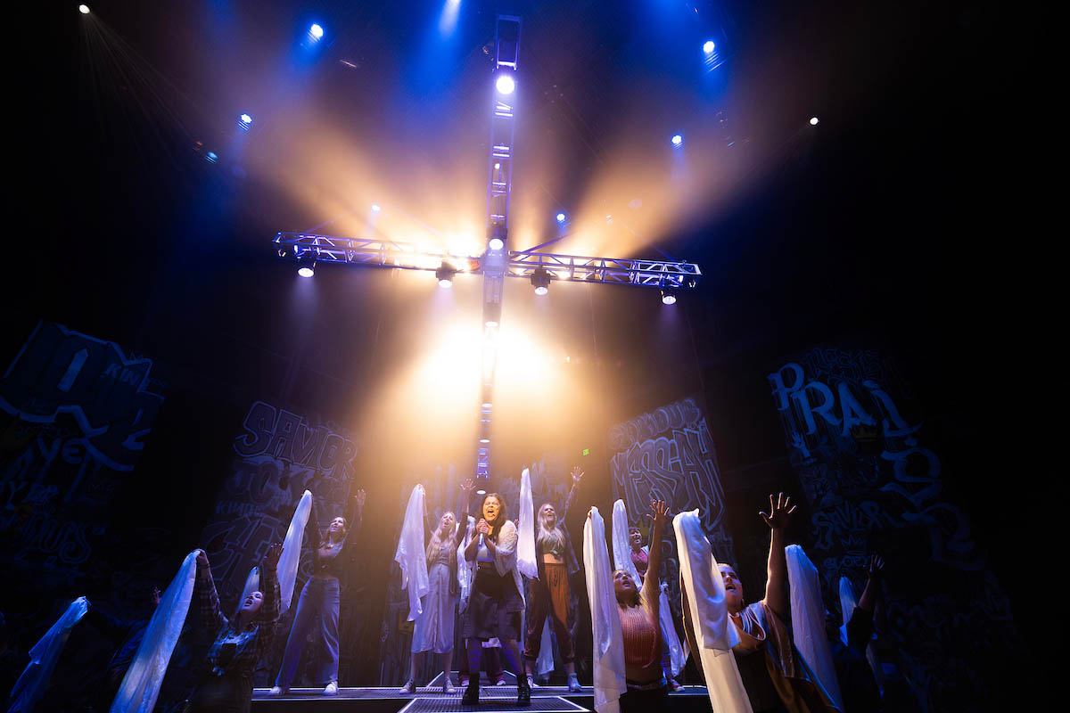 A theatrical performance featuring a central cross illuminated by dramatic lighting. Several performers are on stage, positioned with their arms raised, some holding white fabric. The backdrop includes abstract designs and text, enhancing the visual impact of the scene.
