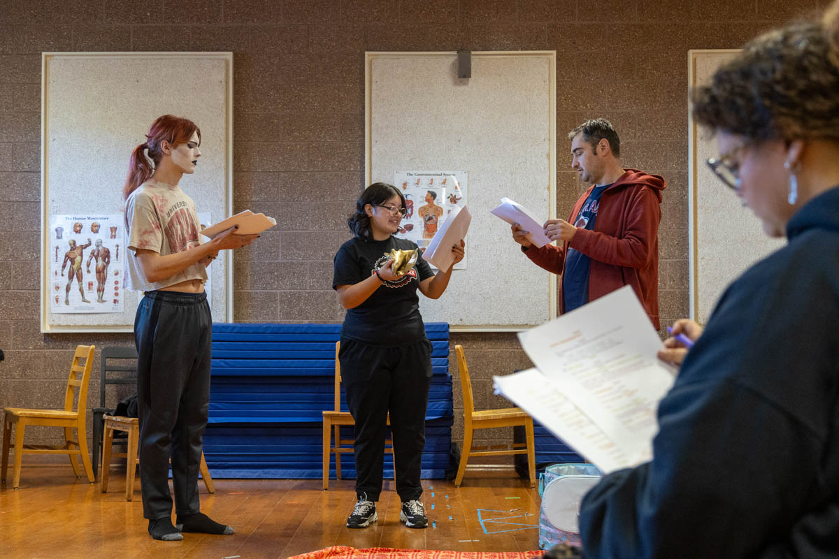 Three people standing in a classroom, reading from scripts; one holds a gold prop mask, while a fourth person takes notes in the foreground.