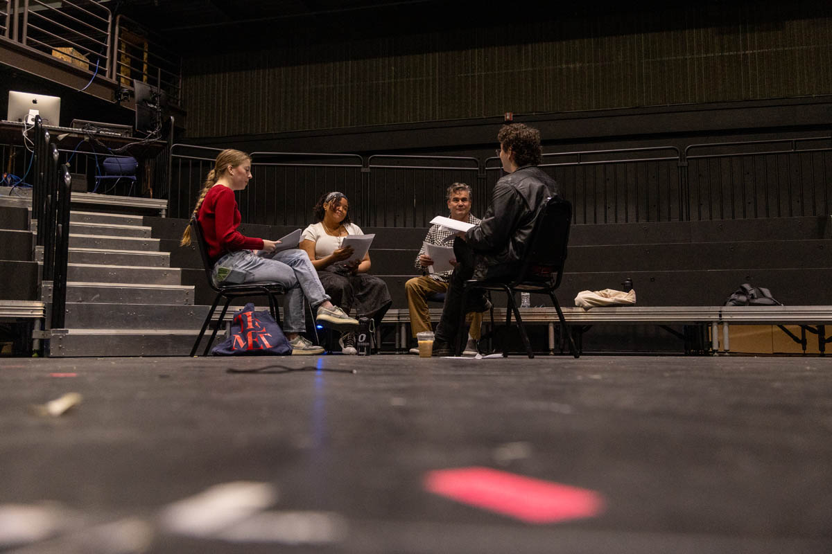 Four people sitting in a circle on a theatre stage, holding scripts during a rehearsal, with empty risers in the background.