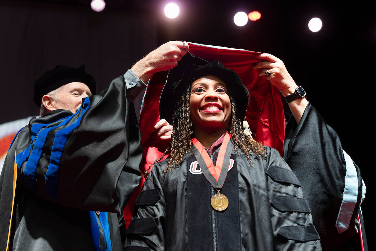 A graduate smiles as a faculty member places a doctoral hood over her shoulders during a commencement ceremony.