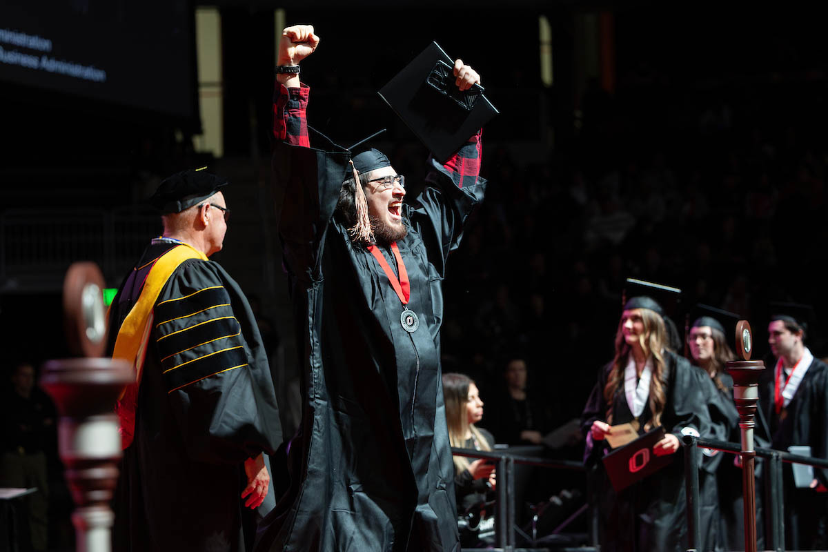 A graduate raises both arms in celebration while holding a diploma cover after crossing the commencement stage.
