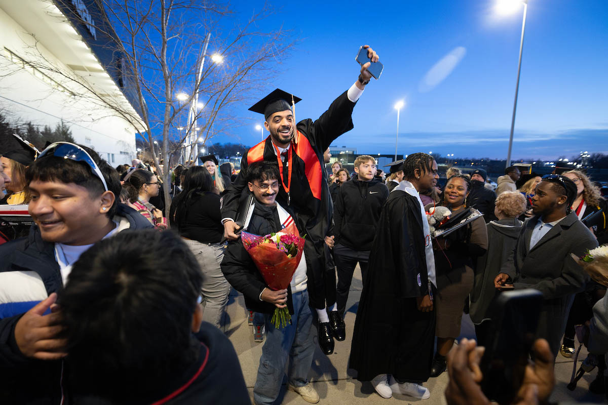 A graduate is lifted onto a friend or family member's shoulders while celebrating with loved ones and fellow graduates outside the venue.