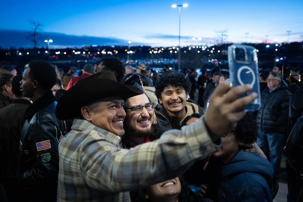 Graduates and family members gather outdoors after commencement, smiling and taking a group selfie at dusk.