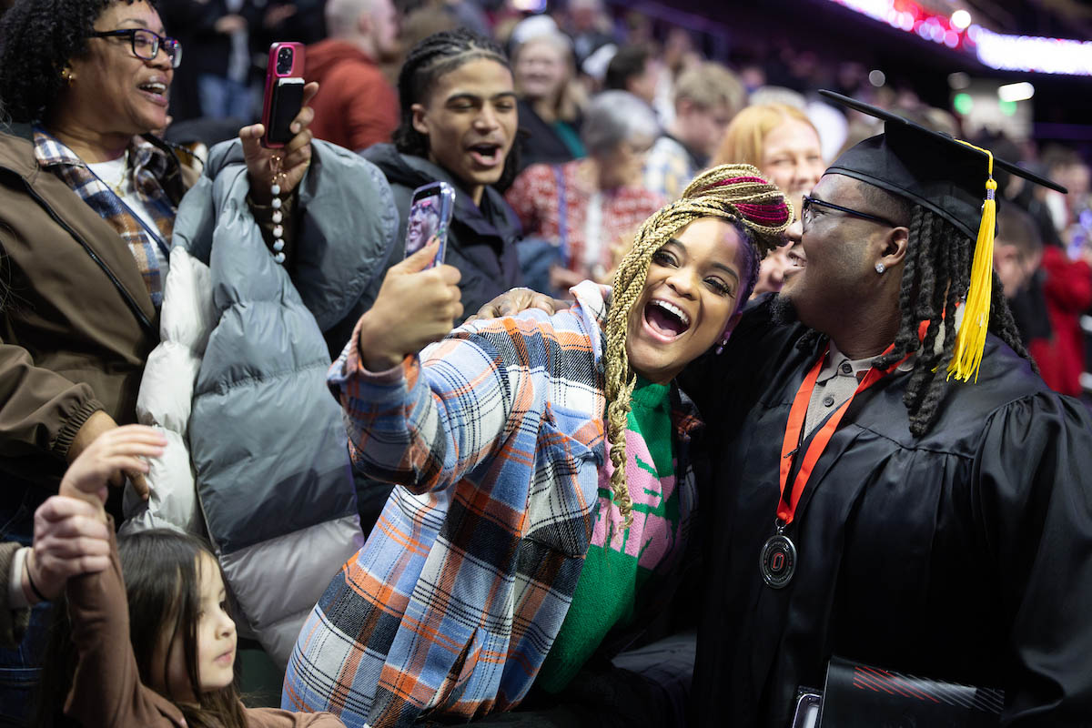 A graduate takes a joyful selfie with a loved one in the stands as others cheer nearby.