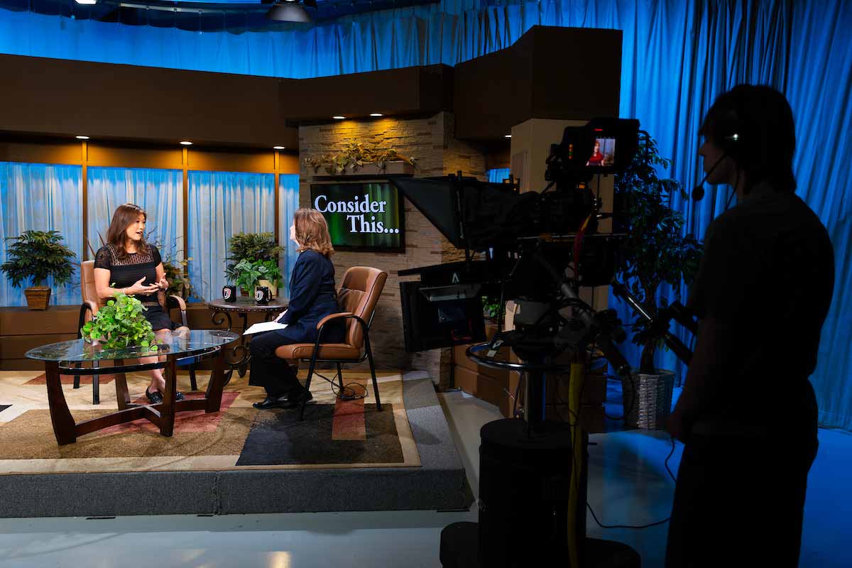 A UNO student stands behind a large camera on a TV set, while two women, Juju Chang and Cathy Wyatt, sit in chairs facing one another in front of a screen that reads ‘Consider This.’ 