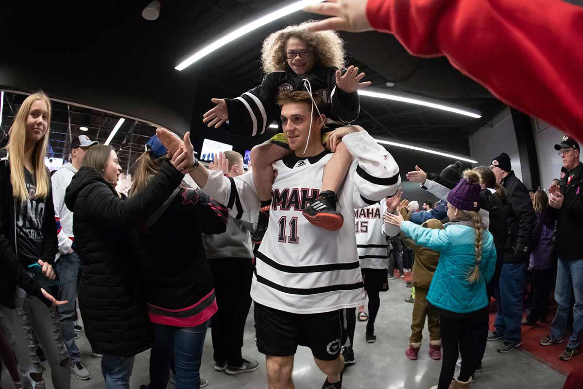 A young boy sits on the shoulders of a hockey player in a white jersey, walking down a hall filled with fans reaching out for high-fives. 