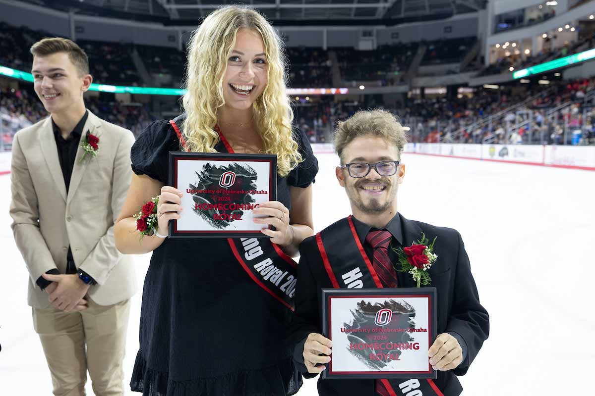 A man and woman wearing sashes pose side by side on the ice at UNO’s Baxter Area, holding plaques that read ‘University of Nebraska Omaha 2024 Homecoming Royal.” 