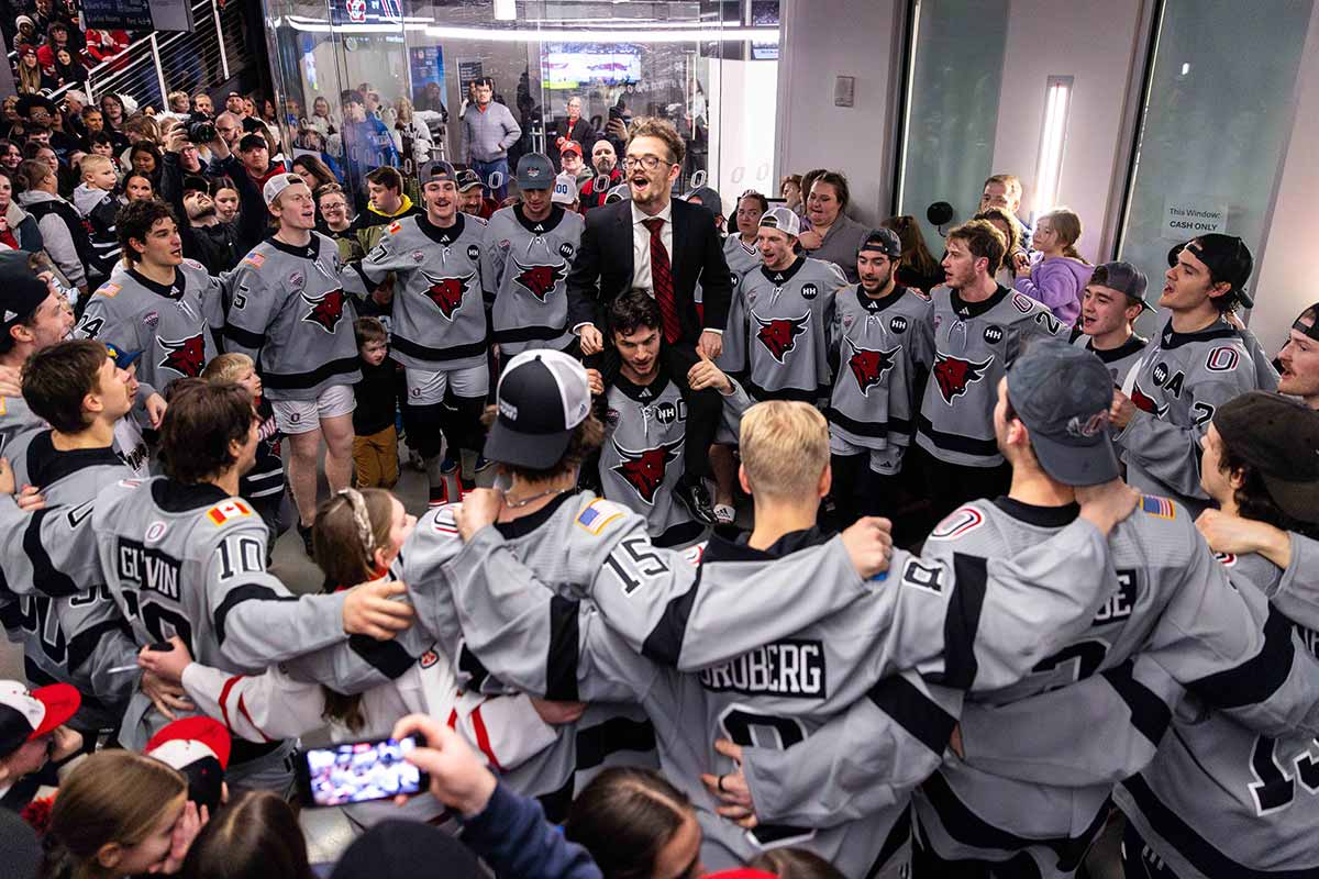 A group of hockey players in gray jerseys stand in a circle singing, while a young man sits on the shoulders of a hockey player in the center of the circle. 