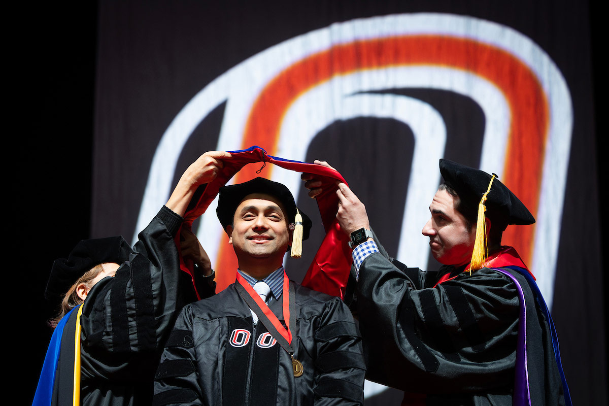 A graduate in black academic regalia smiles on stage as two faculty members place a doctoral hood over his shoulders during a commencement ceremony, with a large red-and-black “O” logo projected in the background. 