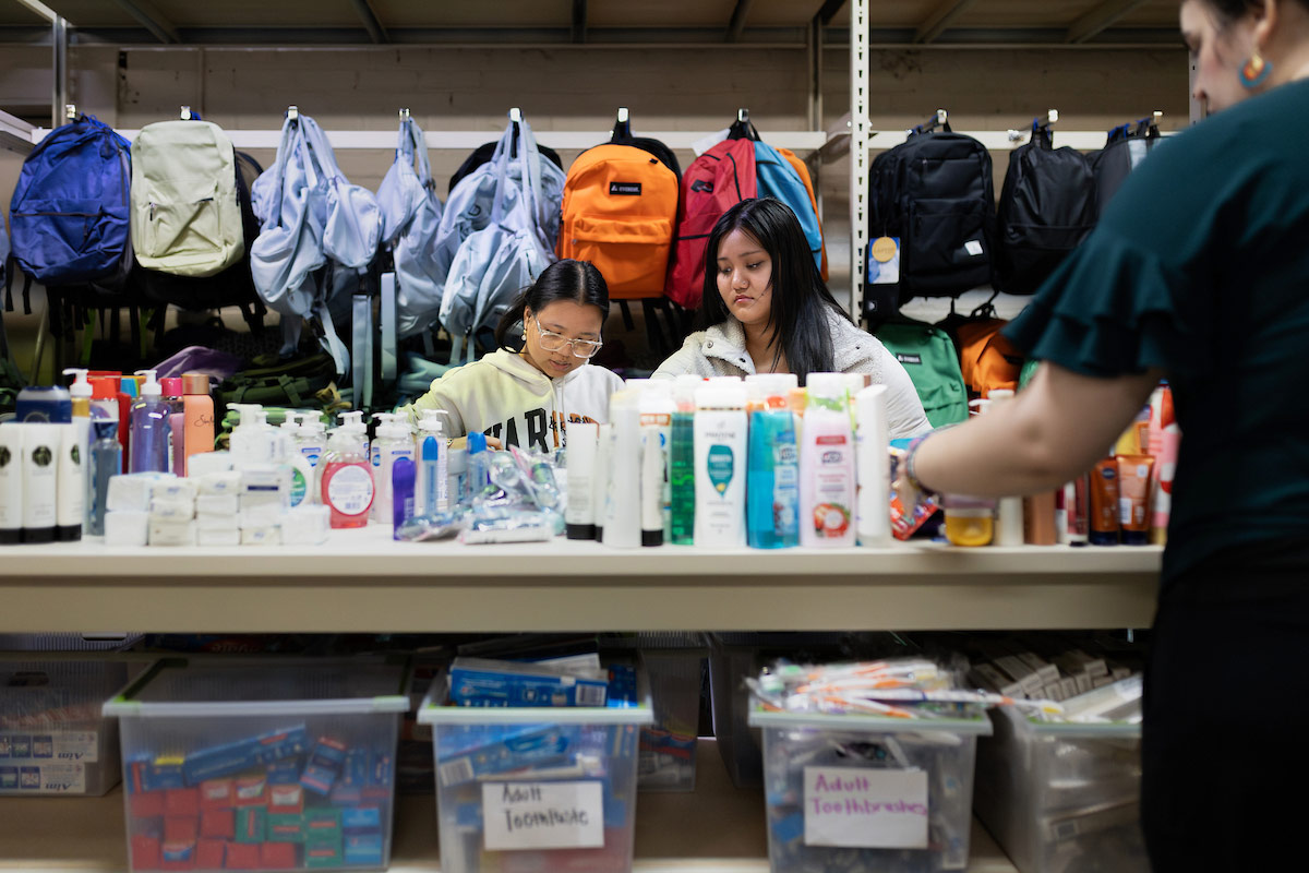 Two students put organize supplies on a table with backpacks arranged behind them.  