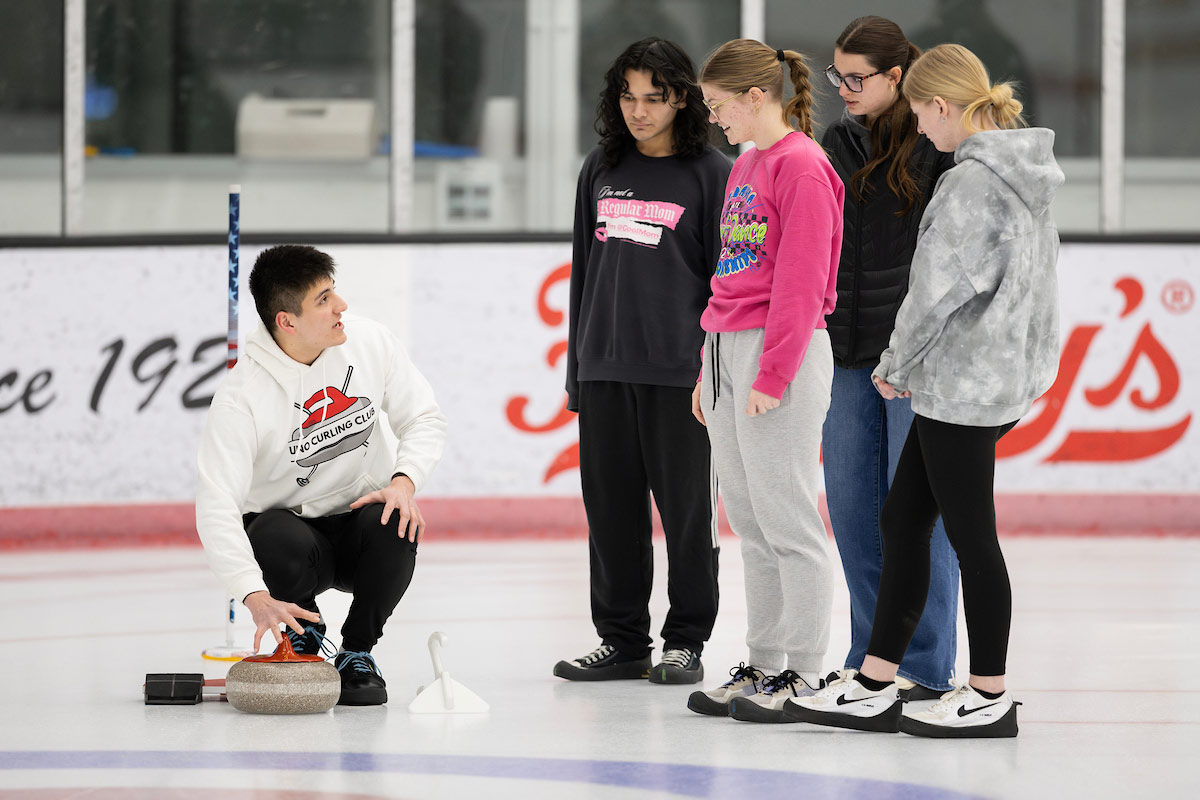 A student kneels over a curling stone and instructs a group on how to curl.