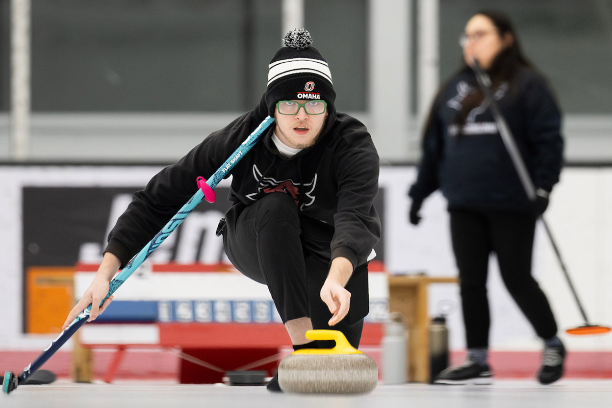 A member of the Curling Club tosses a stone across the ice with a teammate in the background.