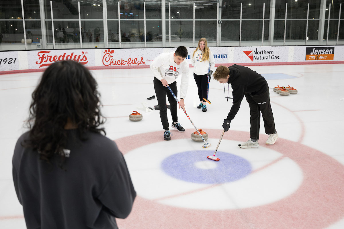 Members of the Curling Team stand on ice and sweep as a curling stone moves across the ice.