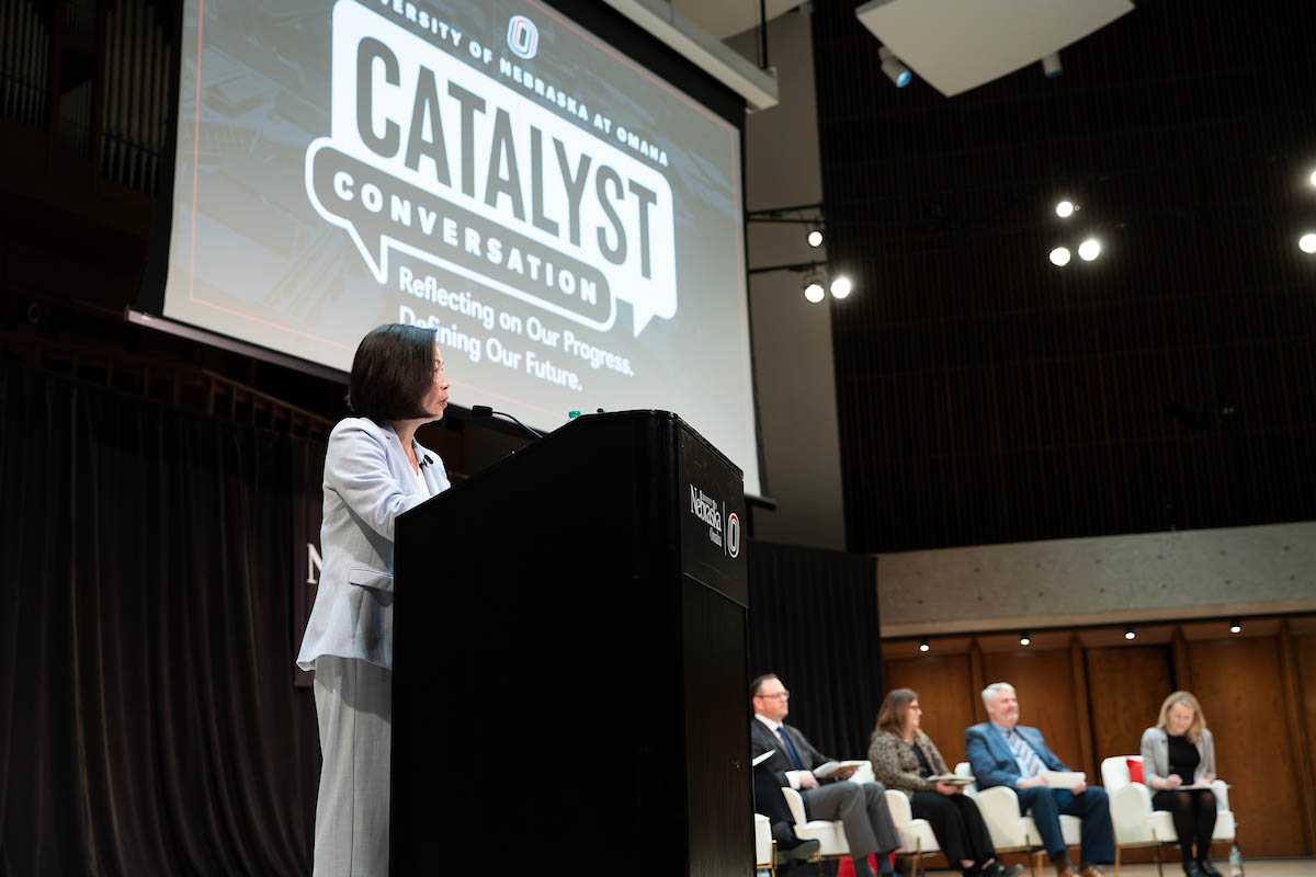 Chancellor Joanne Li stands at a podium speaking to an audience, with panelists seated behind her and a large event screen overhead.