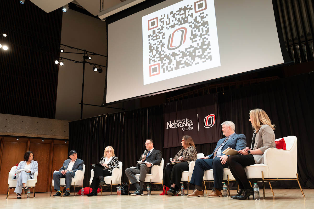 A group of panelists sit onstage beneath a large screen displaying a QR code during the event.