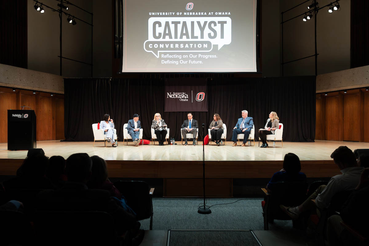 A group of panelists sit onstage beneath a large screen displaying the Catalyst Conversation graphic.