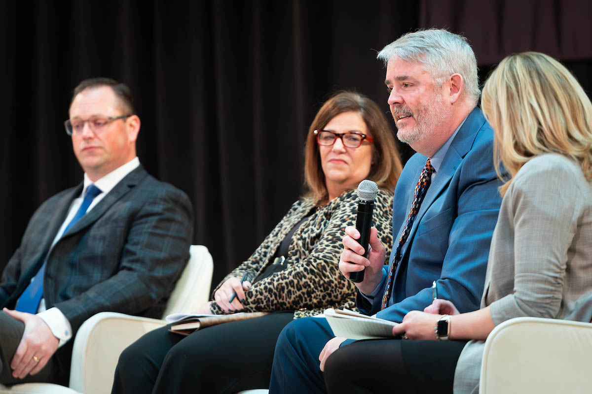 A man speaks into a microphone while seated beside other panelists, including a woman taking notes on the far right.