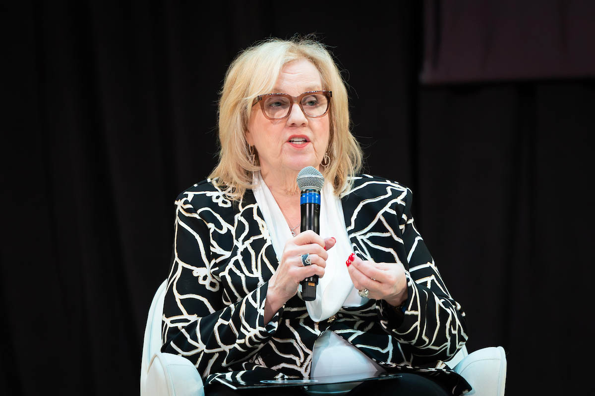 A woman with blonde hair and glasses speaks into a microphone while seated onstage.