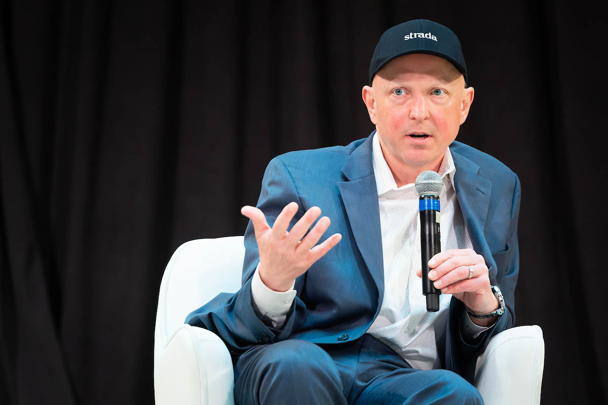 A man in a blue suit and black cap speaks into a microphone while gesturing during a panel discussion.