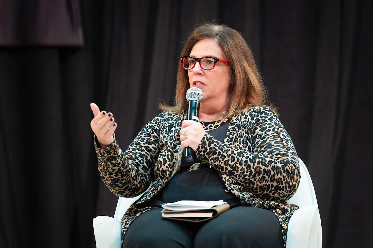 A woman wearing red glasses and a leopard-print jacket speaks into a microphone while seated onstage during the panel discussion.