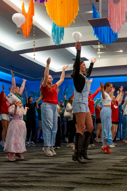 A group of UNO students and Miracle Kids jump up and down with their hands in the air, with colorful decor hanging from the student center ceiling.