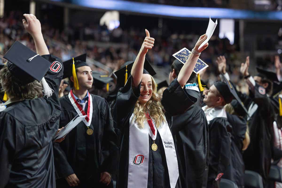 Graduates in black caps and gowns celebrate during a commencement ceremony, raising their hands and smiling.