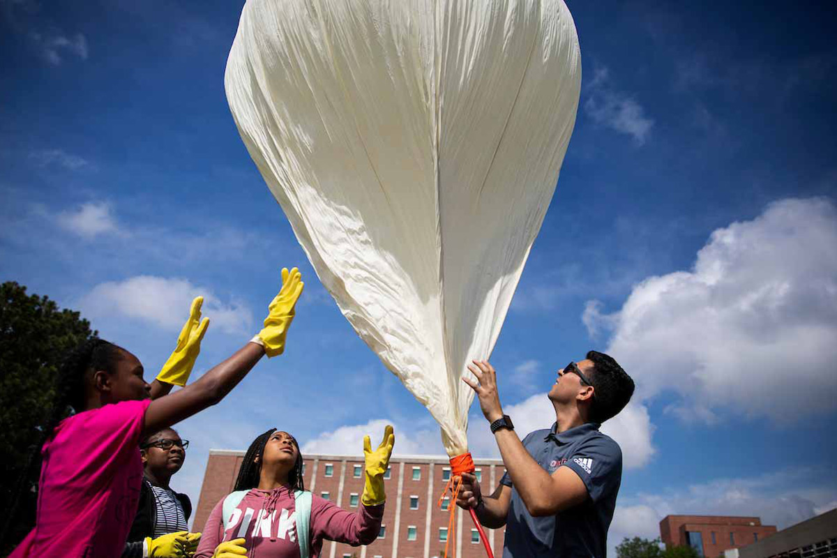A UNO student holds a large white balloon, while three young students watch and offer support with blue skies in the background. 