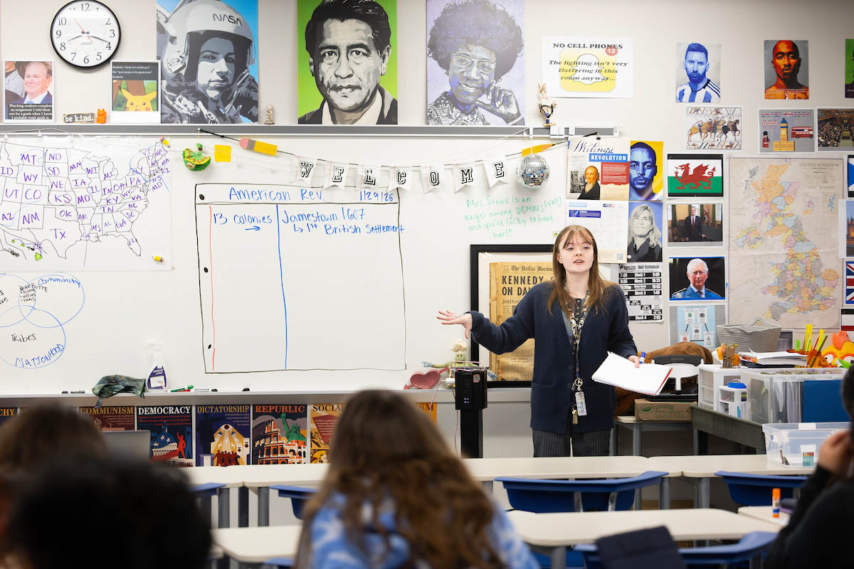 Ellie Sanford stands at the front of a classroom white board with portraits of historical figures above her.