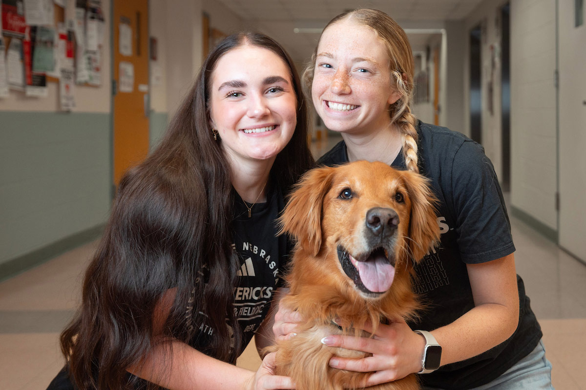 Two UNO students smile and kneel in a school hallway with a happy golden retriever between them, all looking at the camera.