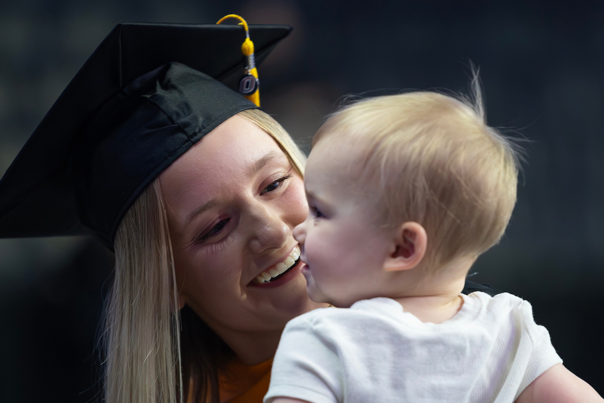 Graduate in cap and gown smiles while holding a baby during the ceremony.