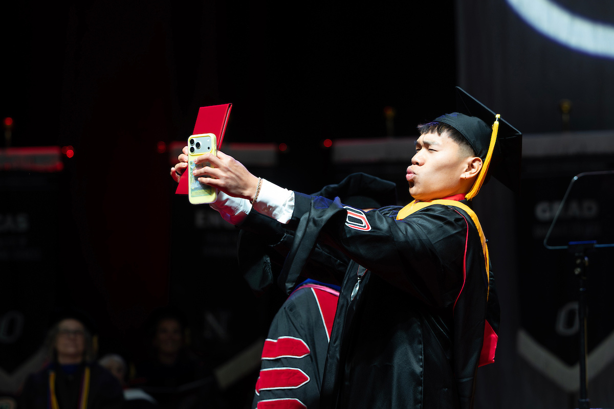 Graduate in regalia takes a selfie on stage while holding a diploma.