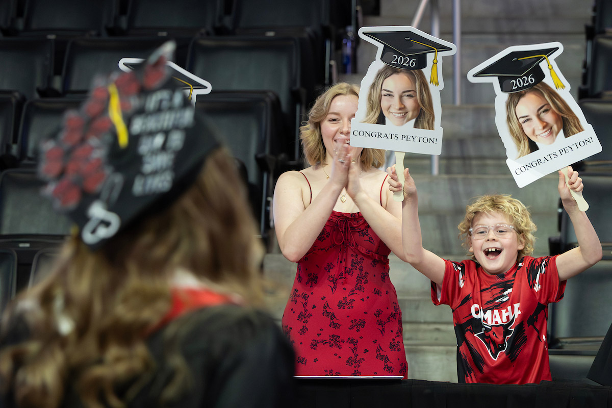 Family members cheer and hold “Congrats Peyton! 2026” signs toward a graduate in cap and gown.