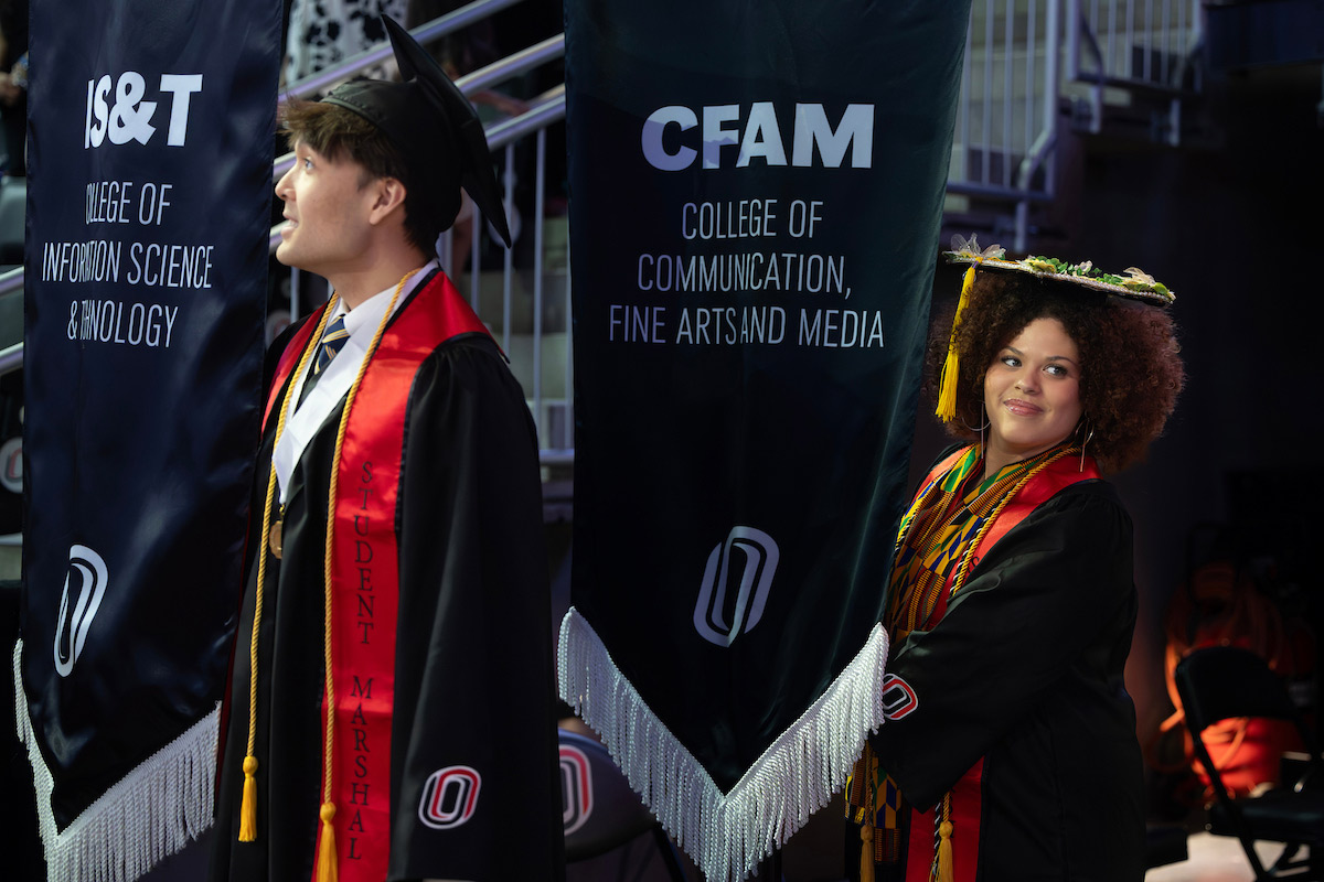 Student marshals in graduation regalia carry college banners during a commencement ceremony.
