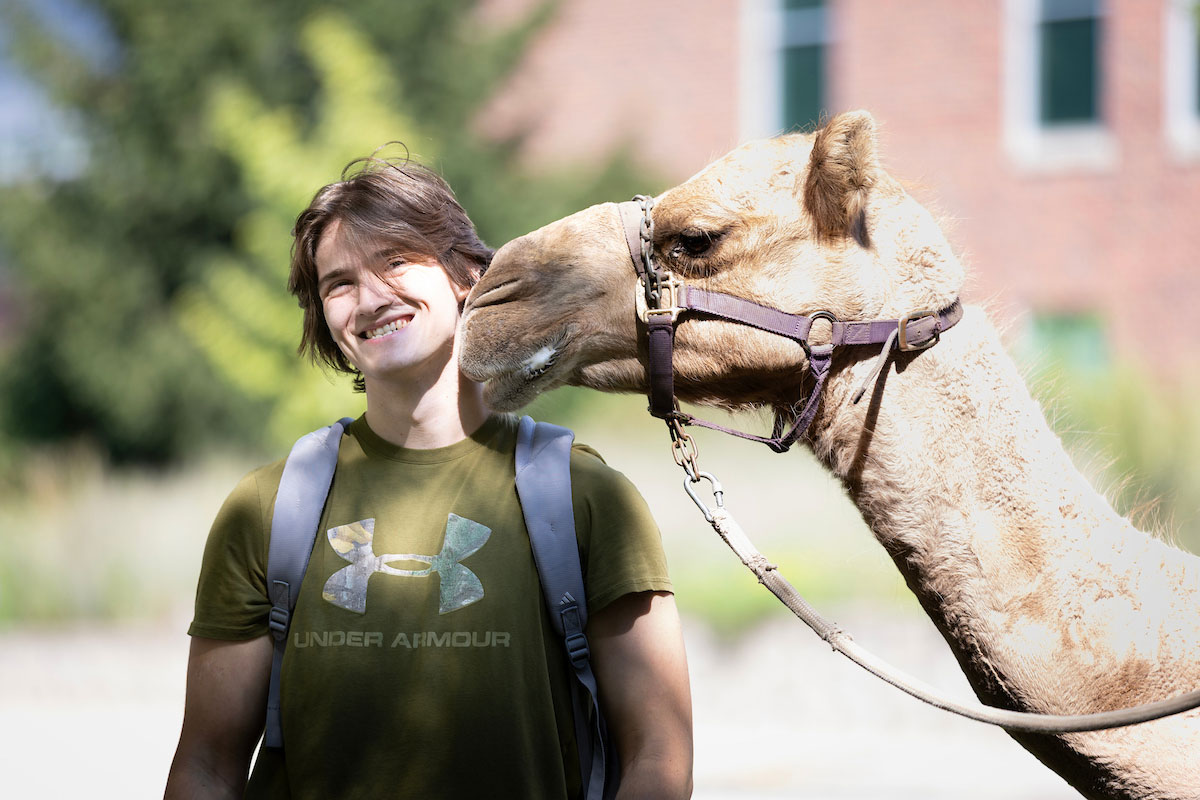 A student smiles and poses while a camel sniffs his face,