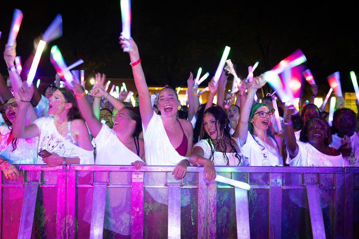 A crowd at a nighttime concert cheers and raises glowing multicolored foam sticks behind a metal barrier, illuminated by vibrant pink, purple, blue, and green lights.
