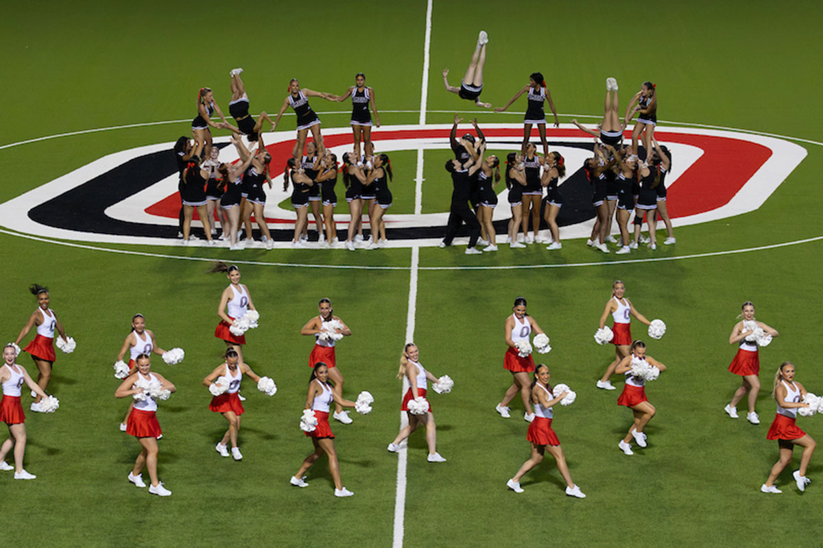 Two groups of cheerleaders perform on a football field, with one group forming a stunt and tossing teammates into the air while another group in red skirts and white tops performs with white pom-poms in the foreground.