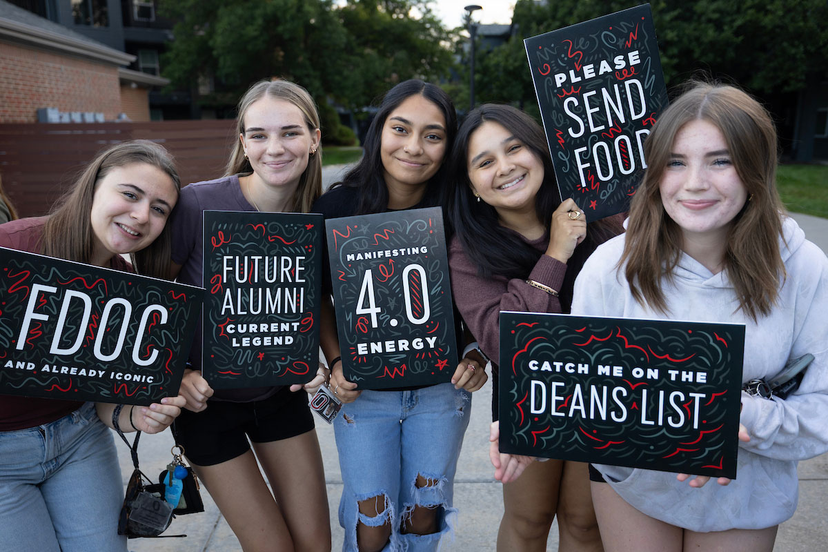 Five students pose outdoors, smiling and holding colorful signs with messages about academics, energy, food, and the dean’s list.
