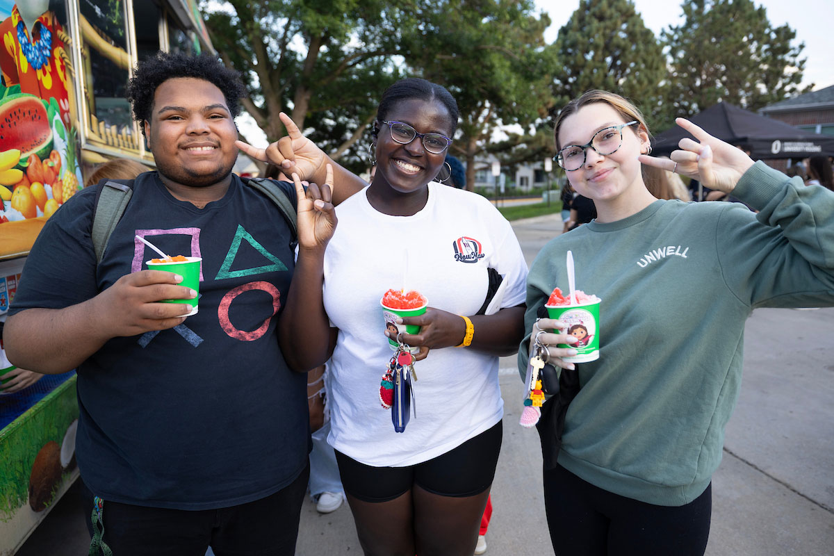 Three friends smile and pose together outdoors while holding cups of shaved ice at a colorful food stand.