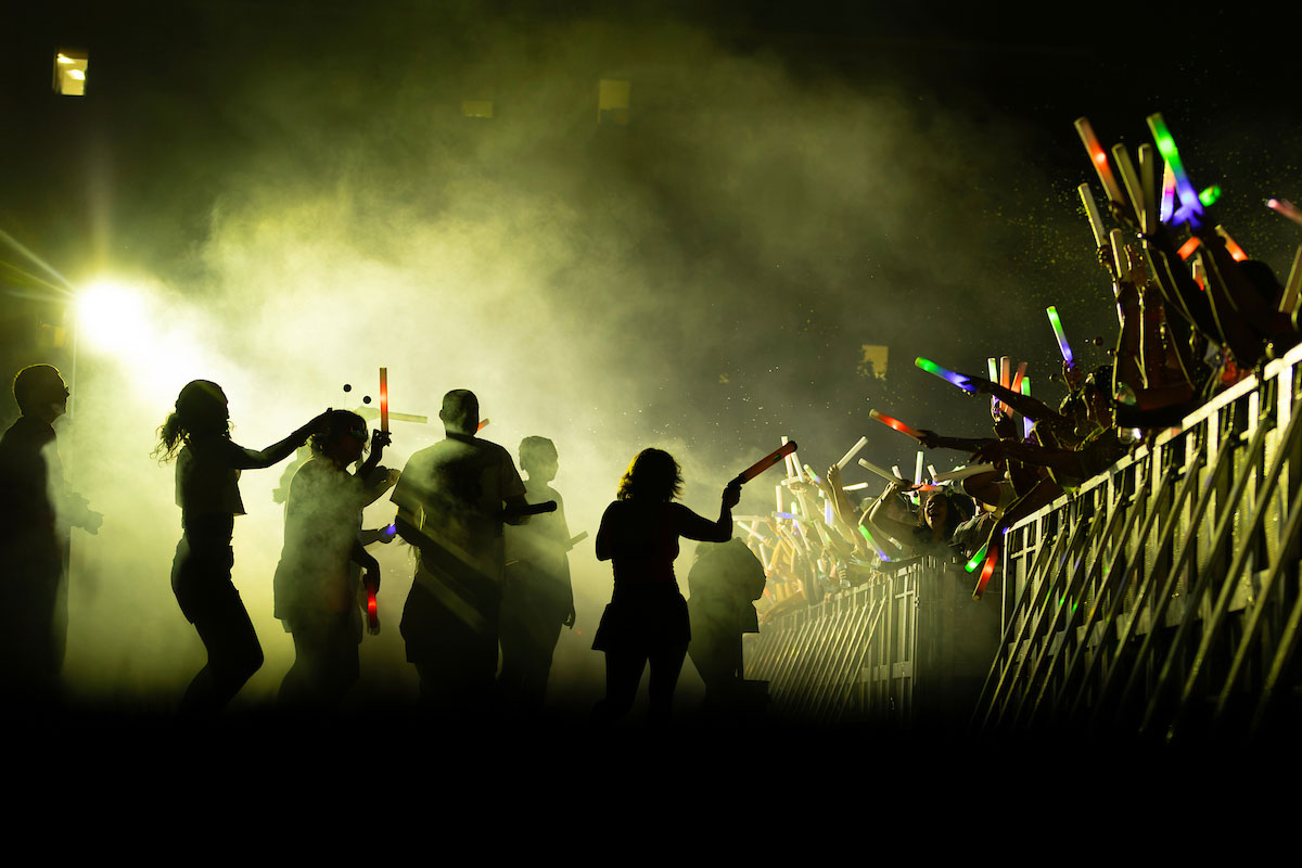 A crowd gathers at a nighttime event, waving colorful glow sticks as silhouetted people move through fog and bright stage lighting.
