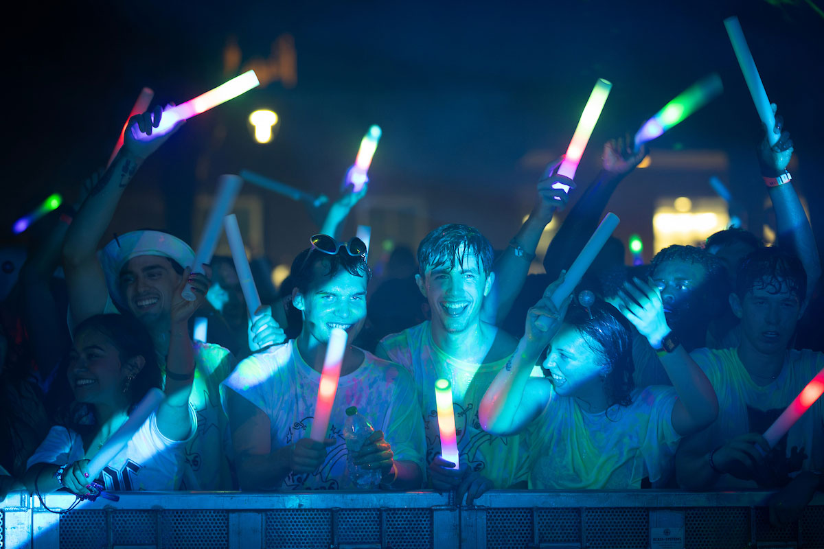 A group of people at a nighttime event smile and raise colorful glowing foam sticks while standing behind a crowd barrier under blue and green lighting.
