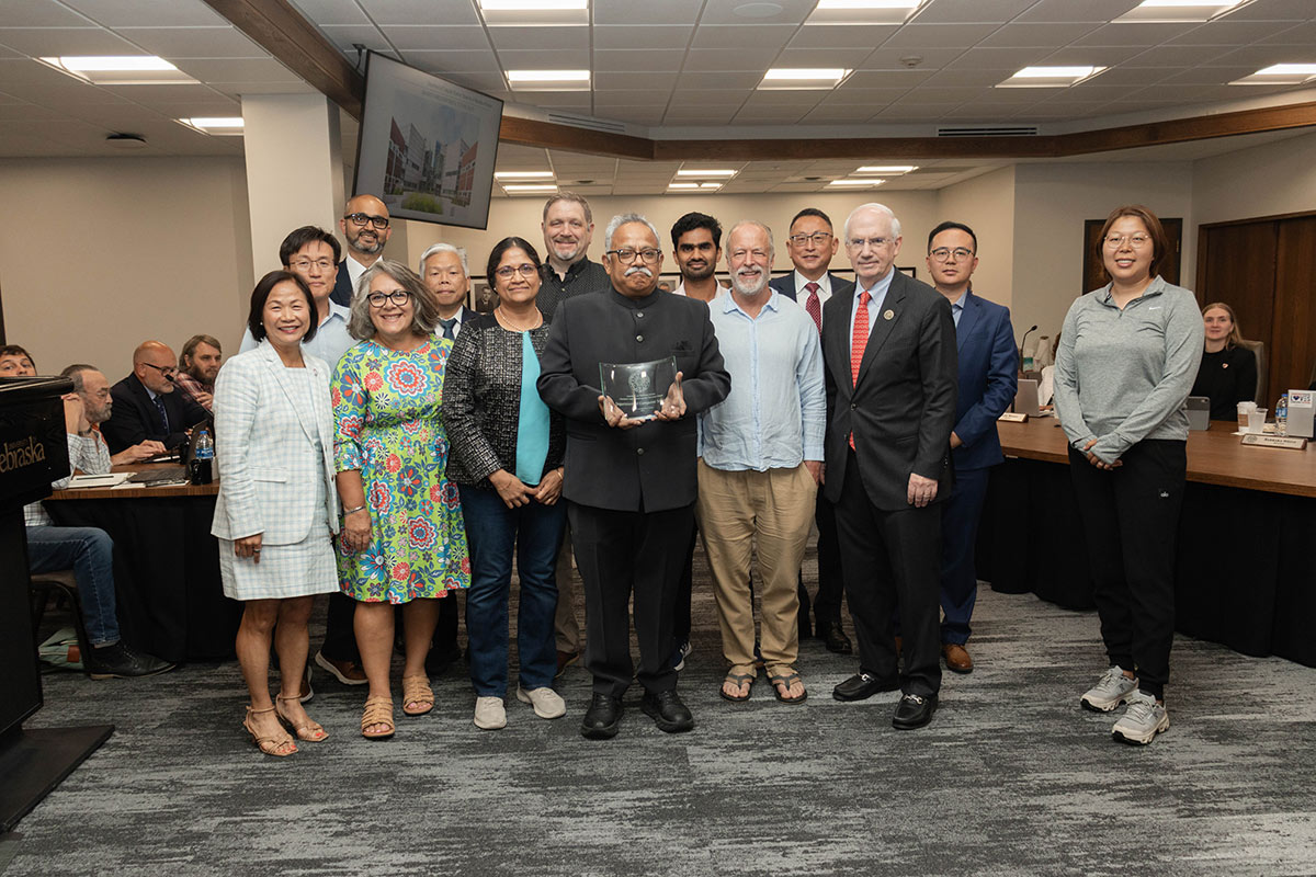 Group photo in a meeting room, with an award recipient holding a glass plaque while colleagues and officials stand around them.