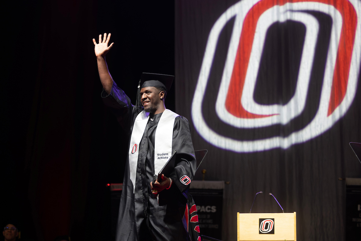 A student wearing a cap and gown waves as he walks across the stage after receiving his diploma. 