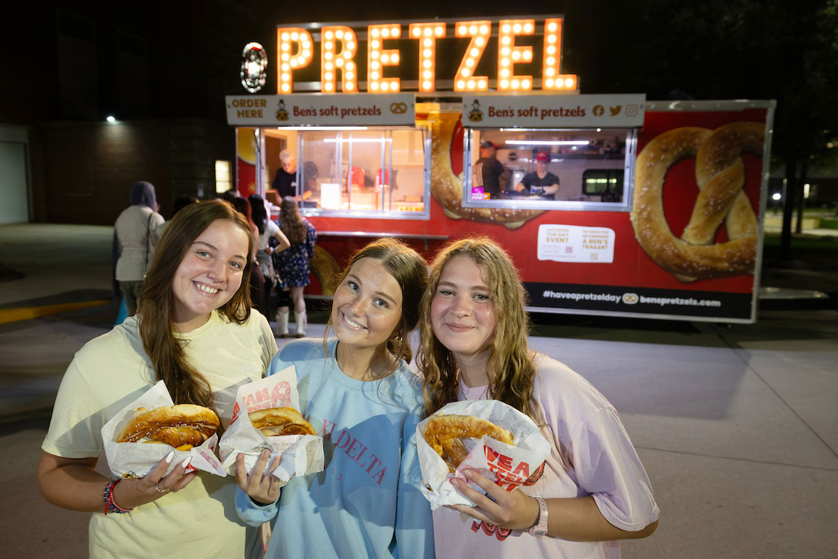 THREE STUDENTS SMILE AND POSE WHILE HOLDING PRETZELS IN FRONT OF A PRETZEL STAND OUTSIDE AT NIGHT.
