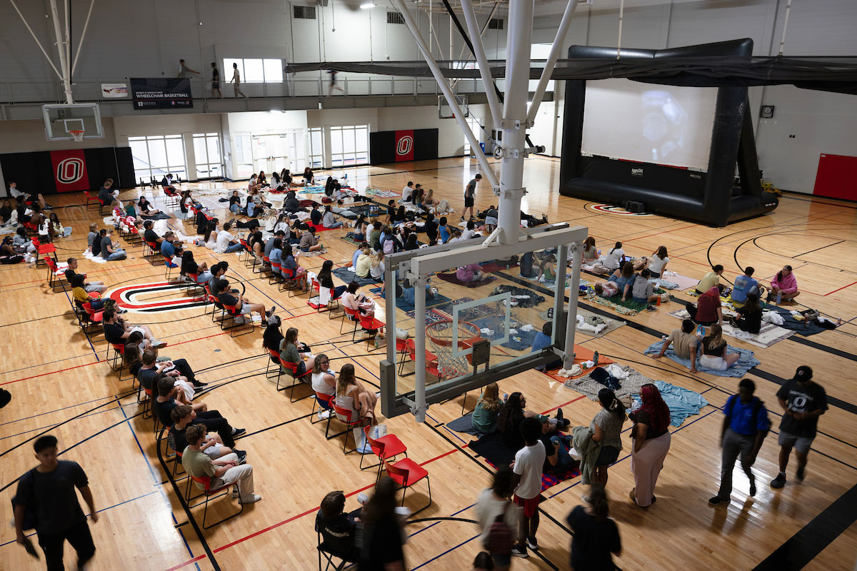 A large group of people gathers on blankets and chairs inside a basketball gym for a movie night, with a large inflatable movie screen set up on the court.
