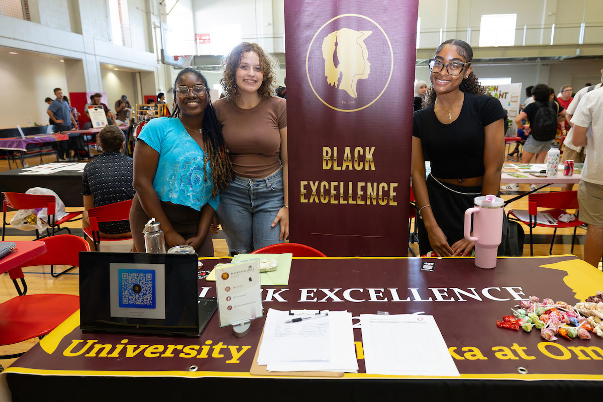 Three students pose behind a table at a campus event, with a large “Black Excellence” banner and university-themed display in front of them. The table features a QR code, informational materials, and candy.
